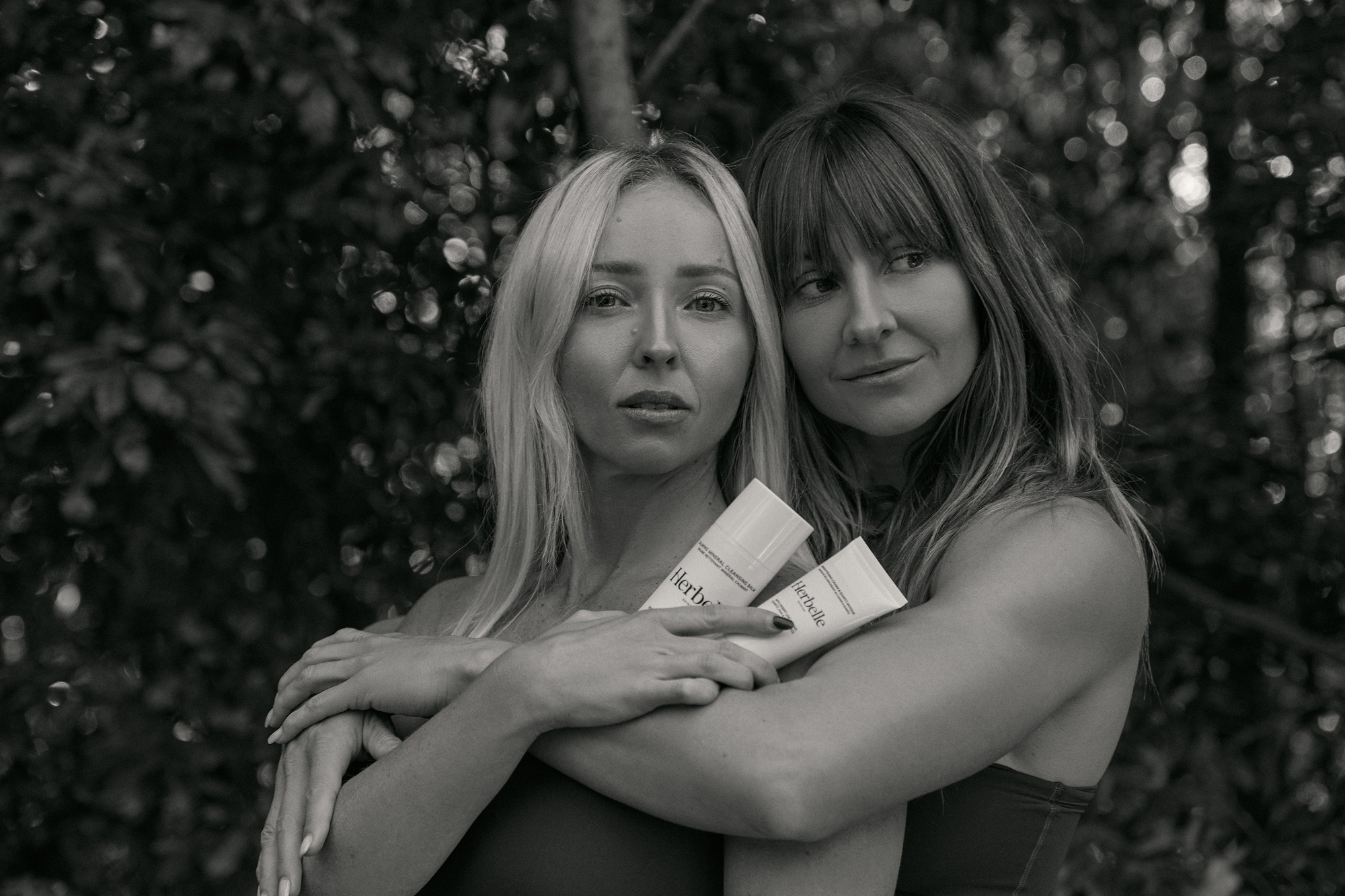 Black and white photo of blonde and brunette females hugging with Herbelle Skincare in hand.