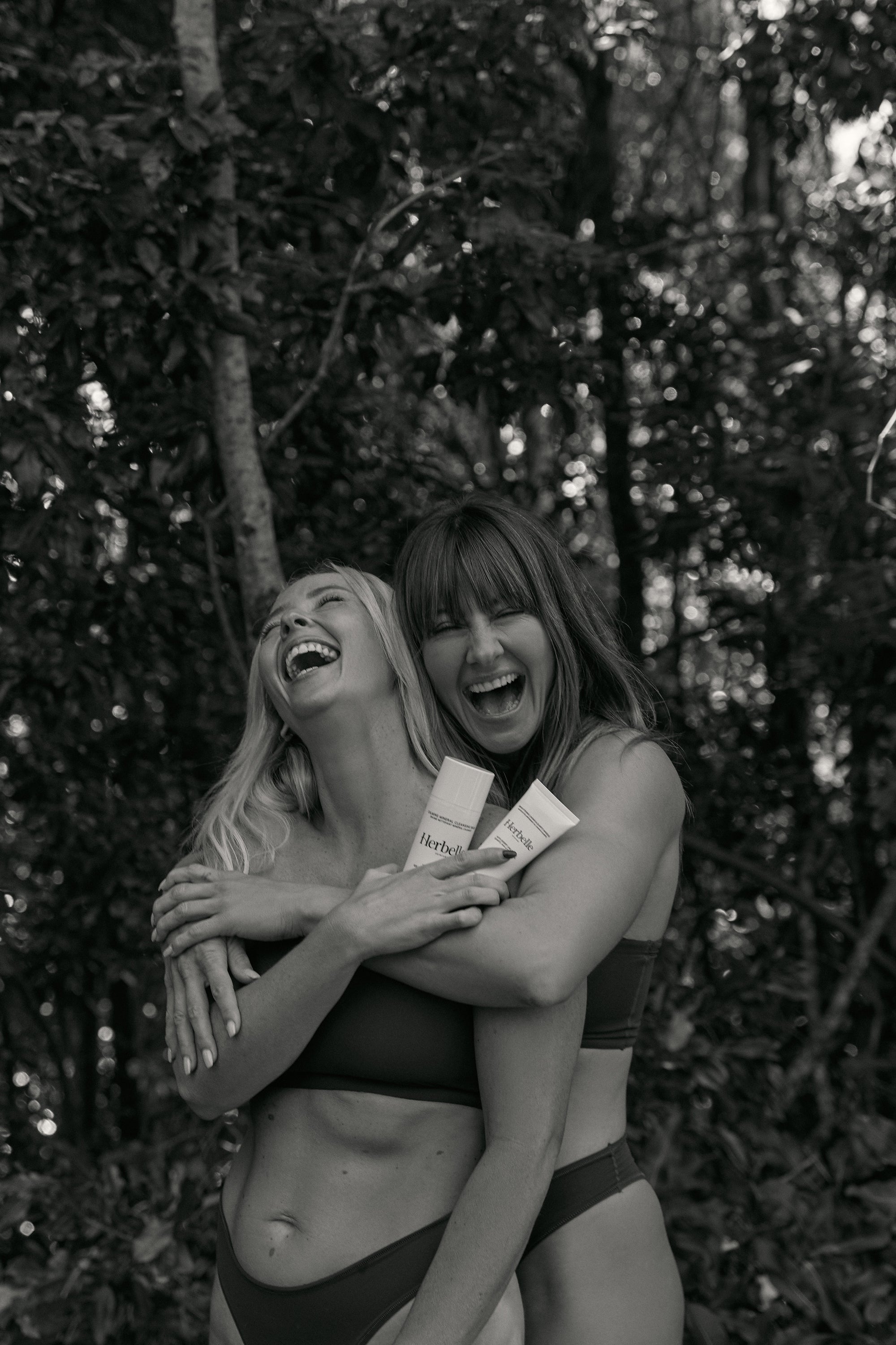 Black and white photo of two women holding each other laughing with Herbelle skincare in hand.